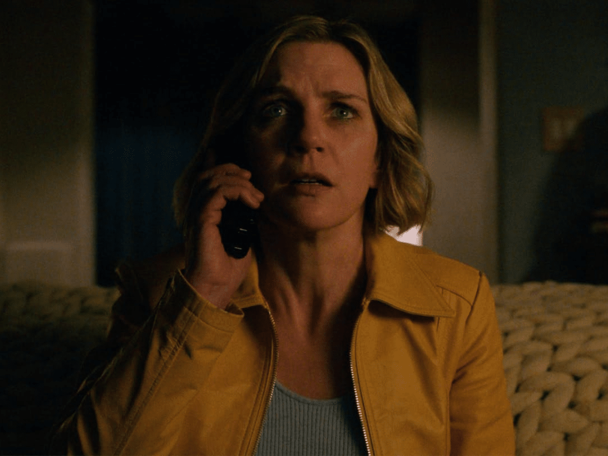 Rhea Seehorn as Carol in Pluribus, wearing a yellow jacket and holding a phone, with a concerned expression.