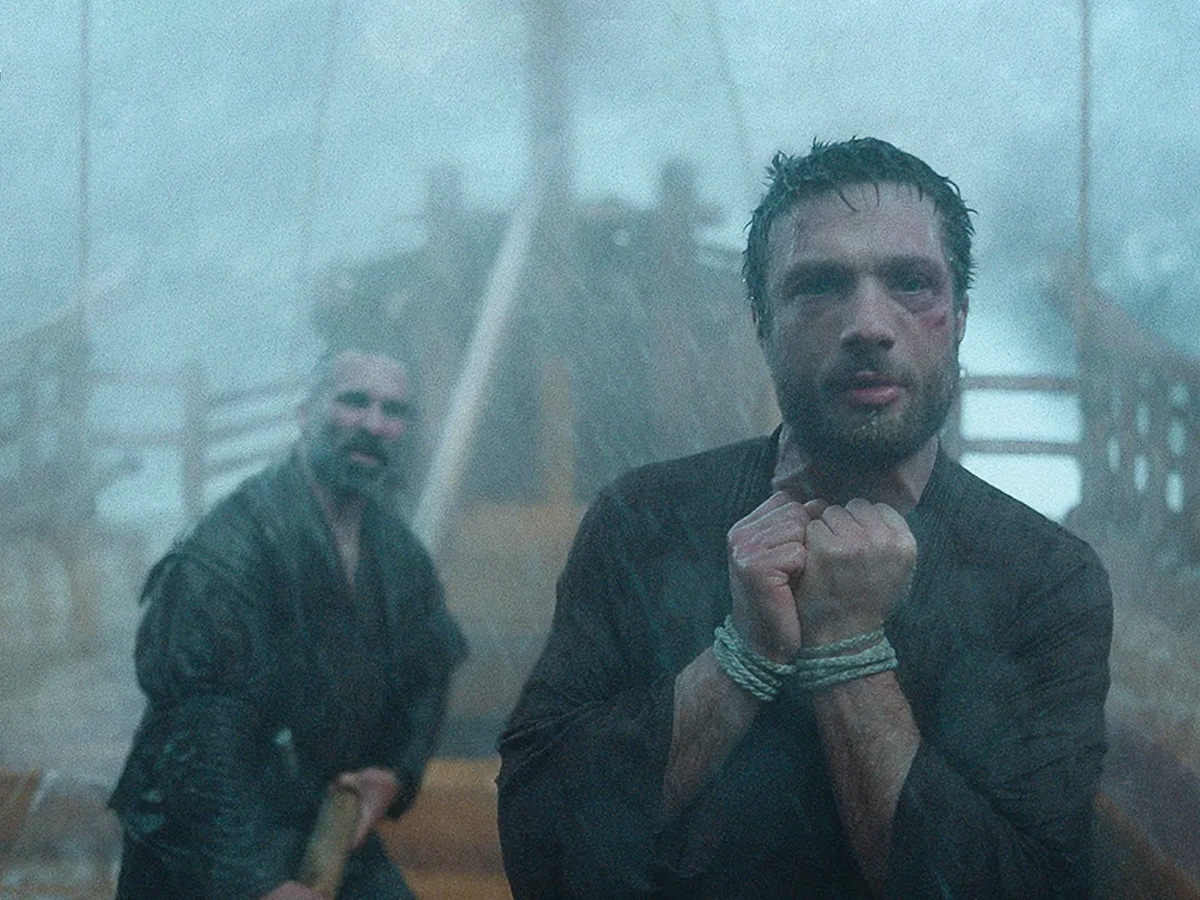 Cosmo Jarvis stands on a ship in a storm, hands bound, wearing a dark robe, with a crew member in the background.