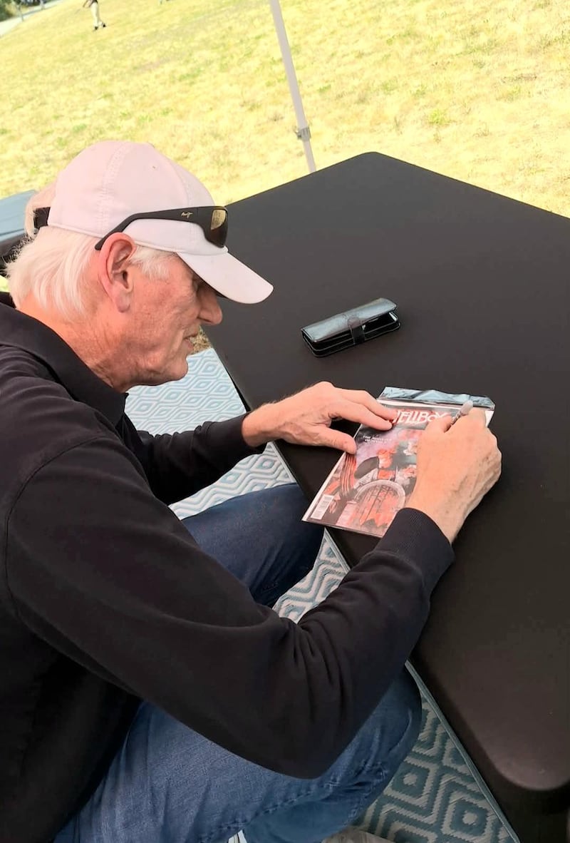 photo taken over shoulder of man in black long sleeve and white baseball cap sitting at a table, signing a wrapped copy of "Hellboy"