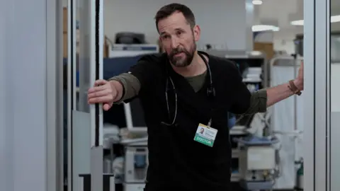 HBO MAX Dr. Michael “Robby” Robinavitch (Noah Wyle) looking exhausted, wearing black scrubs and holding a door open