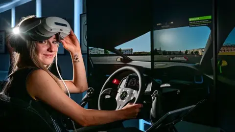 Barnsley College A smiling young woman sits in a driving simulator, in a darkened room,  wearing a virtual reality, she is looking into the camera with the headset lifted above her eyes.