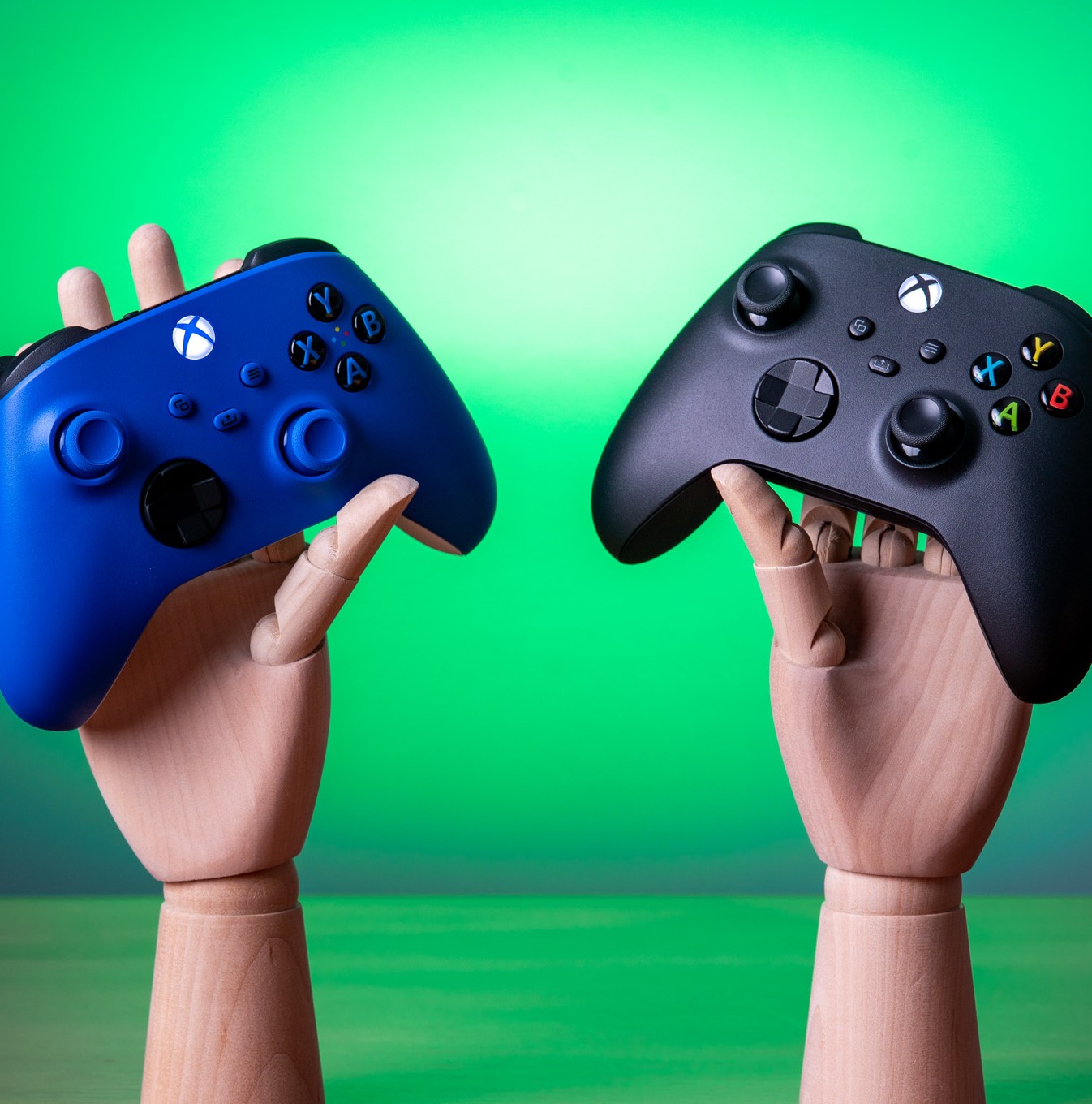 Two Xbox controllers, one blue and one black, being held by wooden mannequin hands.