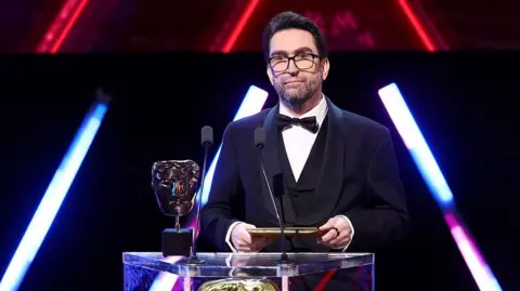 Getty Images A man wearing glasses and a three-piece suit with bow tie stands on-stage behind a lectern, holding an envelope in both hands. On the lectern are two microphones and a Bafta award - a golden trophy shaped like a theatrical mask.