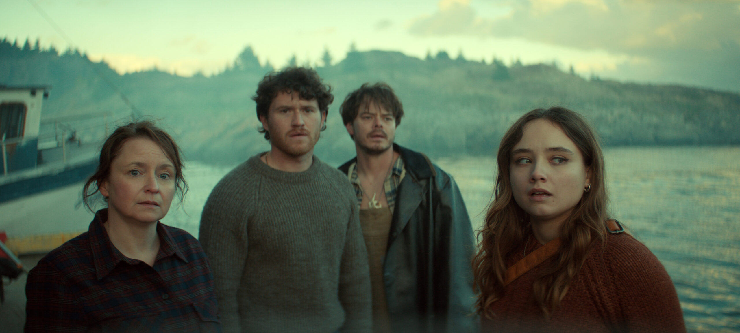 &Quot;Four People Stand Near A Waterfront, Looking Concerned. Behind Them Is A Boat And A Rocky Shoreline With Pine Trees Under A Cloudy Sky. The Lighting Is Moody, Suggesting A Dramatic Or Tense Atmosphere.&Quot;