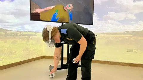 Jessica Bayley/BBC A female student in paramedic uniform stood in a room with projections on the walls. She is wearing a VR headset and a TV screen behind her shows a image of a body.
