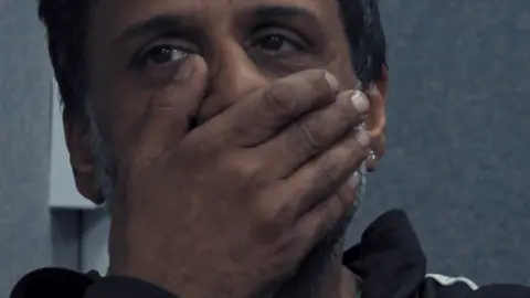 Channel 4 A close-up of the face of Mohammed Hamid, who has his hand clasped to his mouth. His eyes are looking off to his right. Behind him is a grey wall and part of an electricity socket.