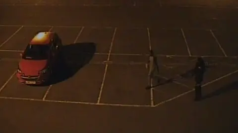 Channel 4 An image from CCTV footage recorded at night. It shows around 10 car park spaces. A red car is parked in one space on the left-hand side of the image while the other spaces are empty. Two people are on the right. One is wearing grey, the other black.