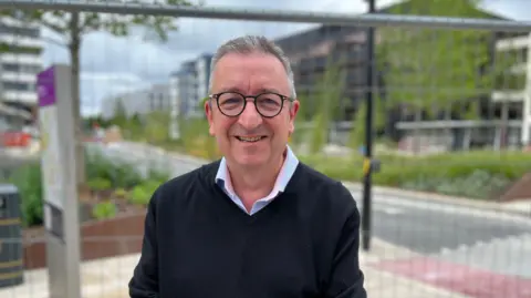 Smiling middle aged man with short straight grey hair and dark rimmed round glasses. He wears a blue and pink shirt, with a black v neck jumper over the top. He is standing in front of some railings with a road and trees in the background.
