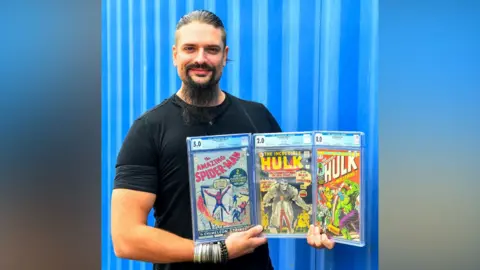 Ewbank's Auctioneers A man holding three comic books against a blue backdrop.