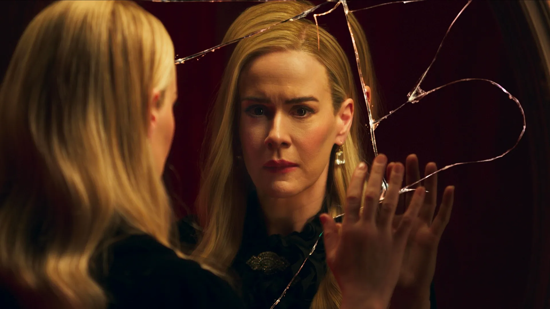 Cordelia Goode (Sarah Paulson) stares into a heavily cracked circular mirror with a distressed expression. Her hand rests gently against the broken glass while her reflection mimics her worried gaze. She wears a black ruffled top accented with a vintage brooch, alongside dangling crystal earrings that contrast against the warm, dimly lit red background.