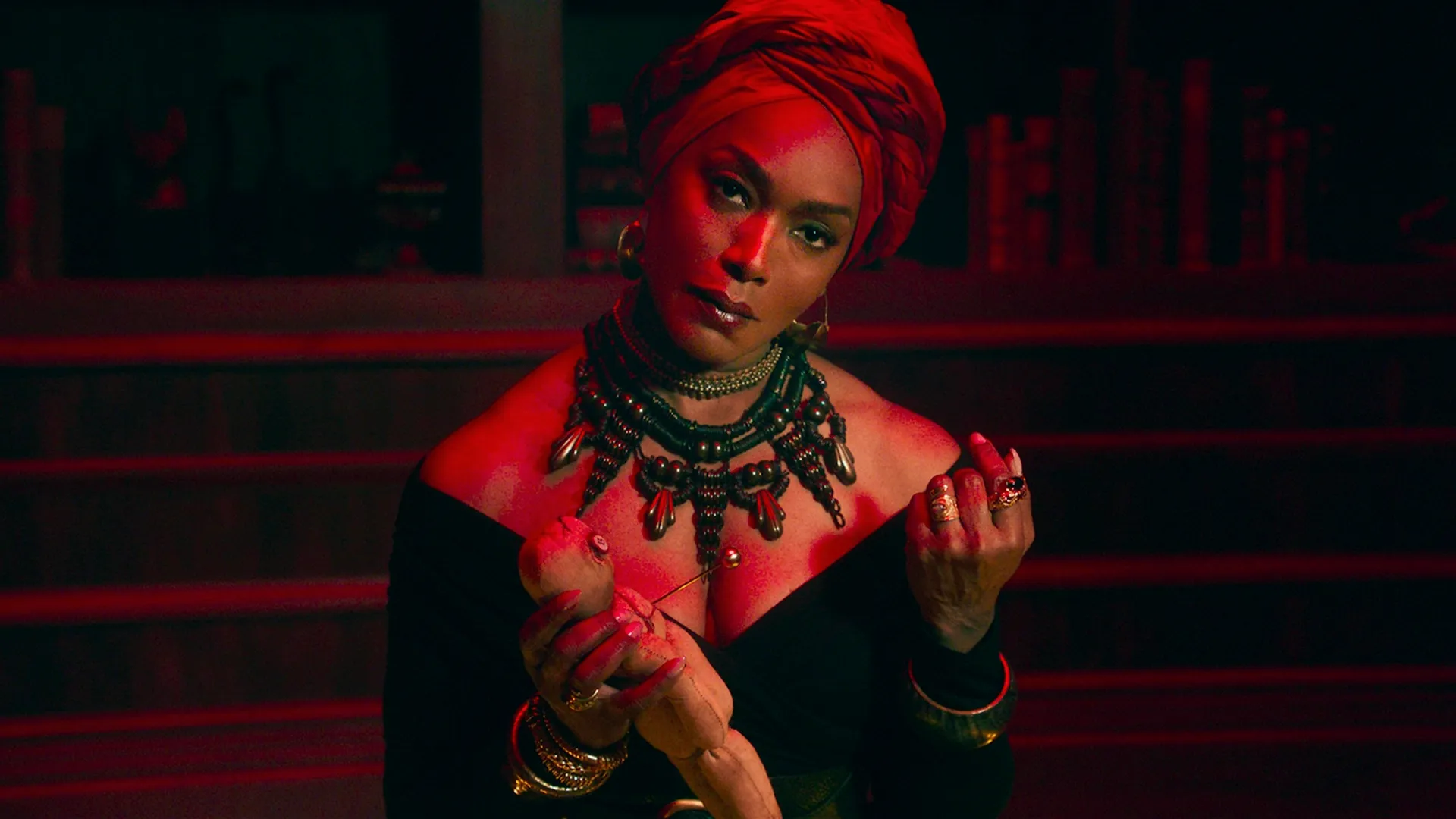Marie Laveau (Angela Bassett) is illuminated by a dramatic red light while wearing a vibrant red head wrap, a heavy statement necklace, and a low-cut black dress as she intensely stares forward, holding a pin above a small doll.