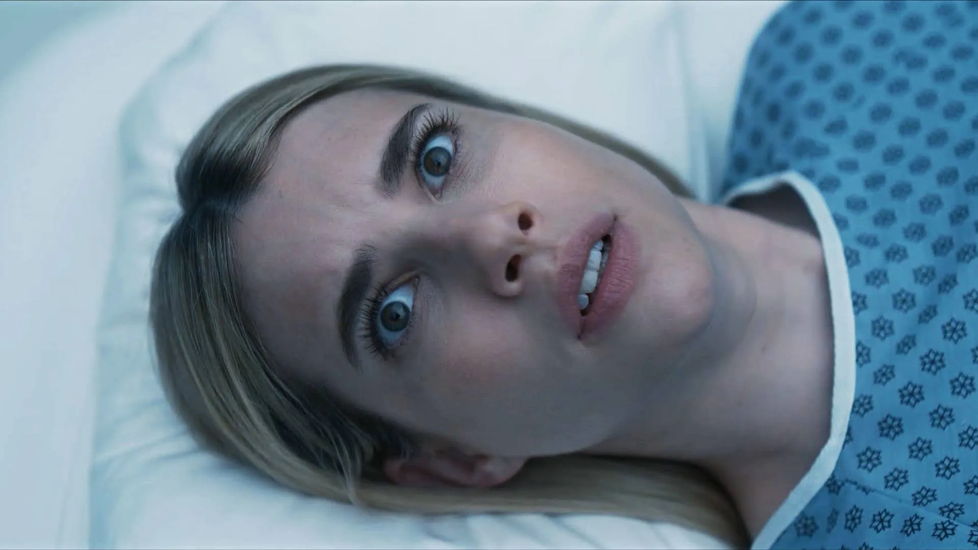 Anna Victoria Alcott (Emma Roberts) lies on her back in a hospital bed, staring wide-eyed and terrified toward the ceiling with a pale complexion and slightly parted lips, wearing a light blue medical gown with a dark blue geometric pattern against a sterile white pillow.
