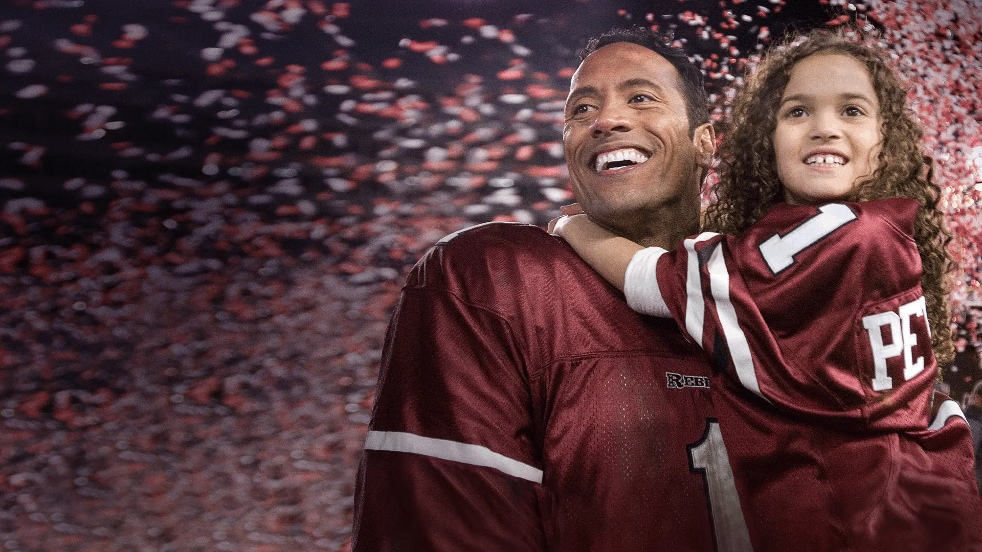 Joe Kingman (Dwayne Johnson), a smiling American football player wearing a maroon jersey, holds a young girl named Peyton Kelly (Madison Pettis) while red and white confetti falls around them in a scene from the Disney comedy movie The Game Plan.