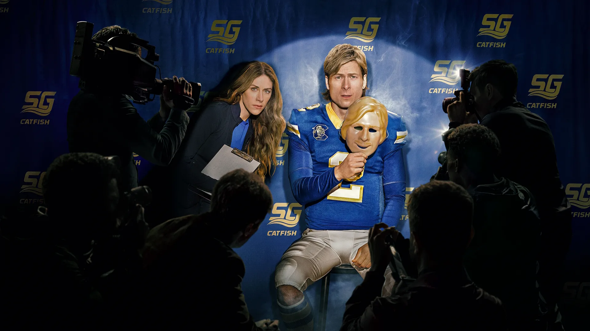 Russ Holliday (Glen Powell) sits under a spotlight during a press conference while holding a mask of his own face, surrounded by cameras and photographers