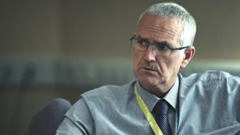 Channel 4 Tom Hamm is on the right hand side of the picture. He is looking off to his right. He has grey short hair and is wearing black-rimmed glasses, pin-striped blue shirt and a lanyard. The background is blurred but he appears to be sitting in an office.
