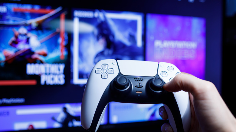 Person holding PlayStation 5 DualSense controller with blurry TV screen in background