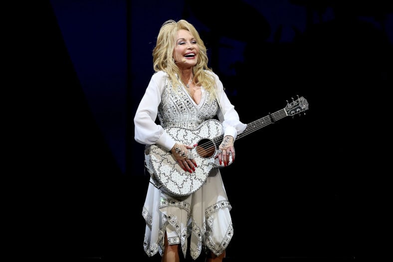 Dolly Parton standing on stage smiling as she plays a white acoustic guitar adorned with sparkling embellishments, wearing a matching white, bedazzled dress with long semi-sheer sleeves.