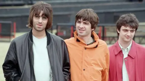 Getty Images The Gallagher brothers together with their band at Knebworth in 1996. Liam has on a black leather jacket and round sunglasses while Noel is wearing an orange cagoule