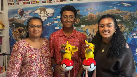 BBC/Emily Johnson Two women and a teenage boy stand indoors in front of a colourful world‑map mural, with two of them holding Pikachu trophies, surrounded by shelves of books and Pokémon items.