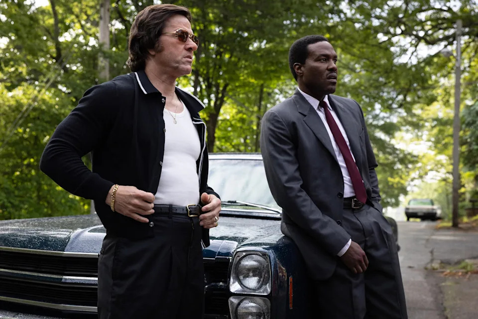 Mark Wahlberg and Yahya Abdul-Mateen II in "By Any Means." Two men stand beside a vintage car on a tree-lined street, one wearing sunglasses and the other a dark suit and red tie.