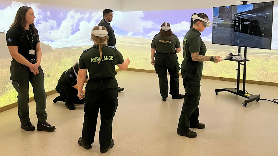 A group of students in paramedic uniform in a room with projections on the walls. They are wearing VR headset and on a TV screen in the room you can see something which looks like a video game.