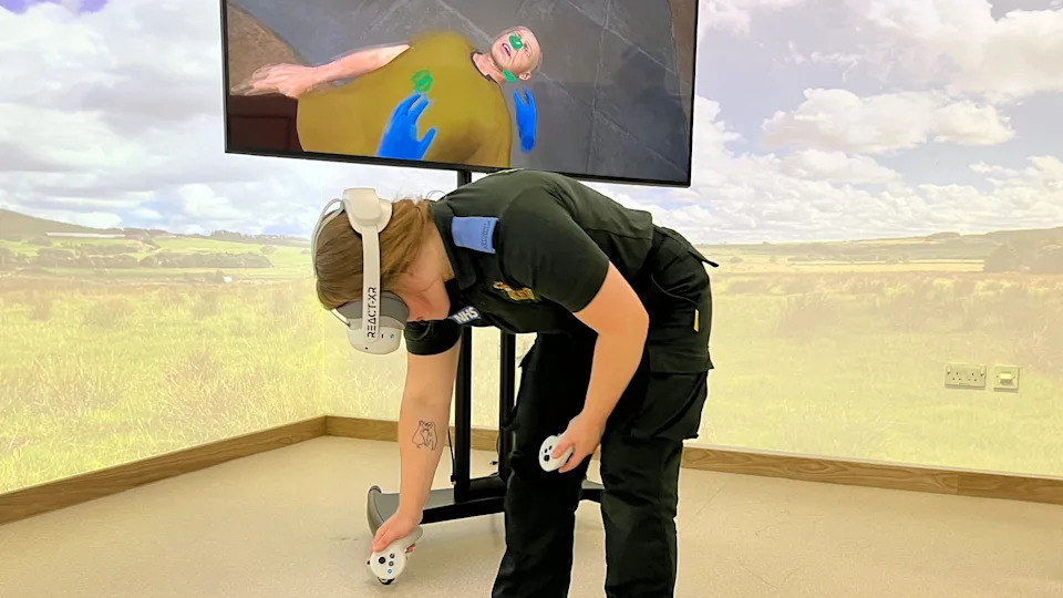 A female student in paramedic uniform stood in a room with projections on the walls. She is wearing a VR headset and a TV screen behind her shows a image of a body.