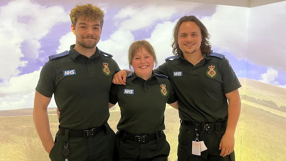 A group of students in paramedic uniform stood together facing the camera. Two boys either side of a girl, there are projections of a landscape on the walls.