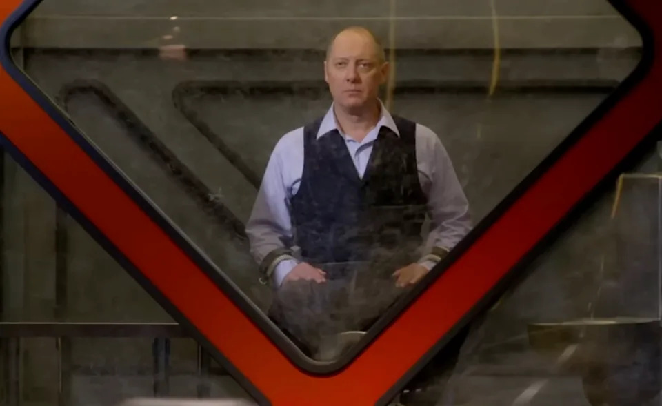 Raymond Reddington (James Spader) is seated behind glass, wearing a formal vest over a shirt, looking forward in a composed manner, within a geometric frame