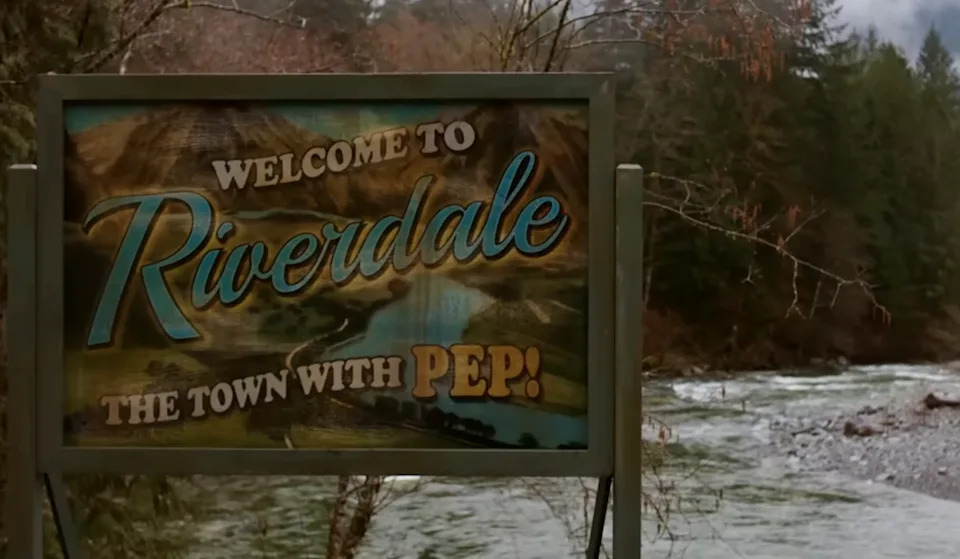Sign reads: "Welcome to Riverdale, the town with pep!" displayed near a river and forest backdrop