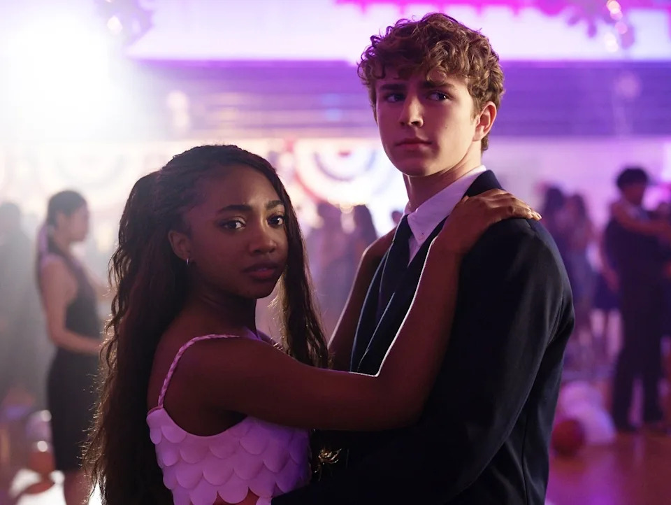 Two teenagers in formal attire dance closely at a school event, surrounded by softly-lit background and other couples