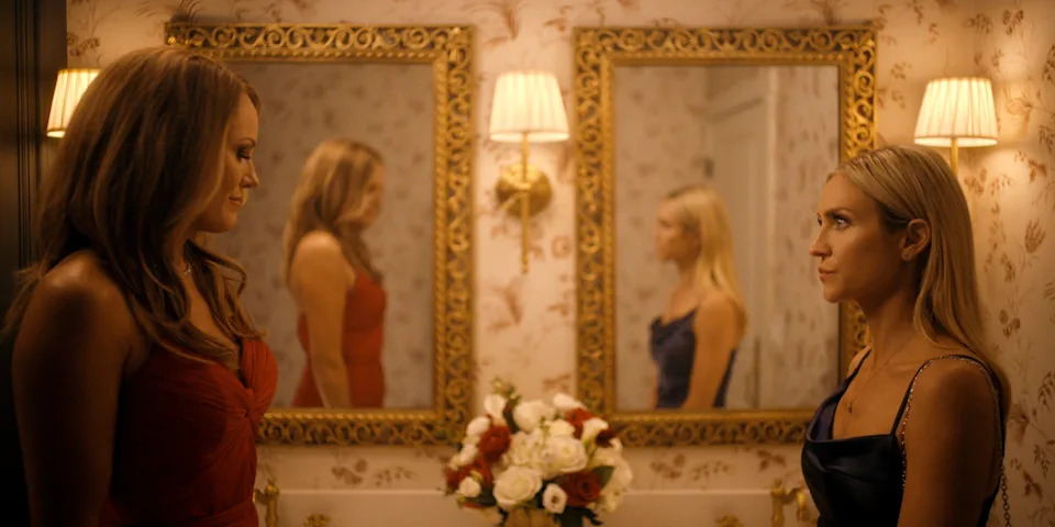 Two women in evening gowns face each other in a luxurious room with floral wallpaper and ornate mirrors