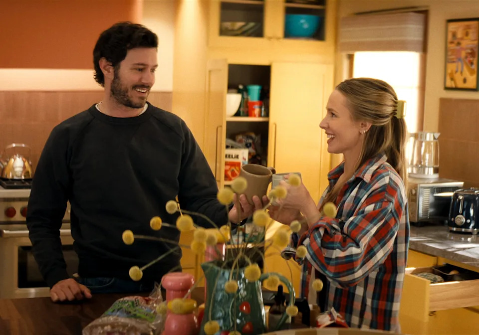 Two people in a casual, homey kitchen share a laugh. One holds a mug, and both appear relaxed and happy