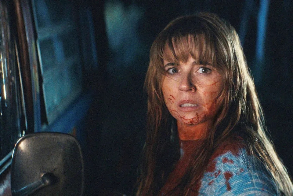 A person with a fearful expression, covered in what appears to be fake blood, stands next to a vehicle, suggesting a dramatic movie scene