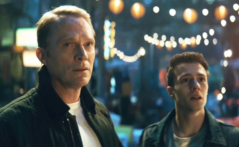 Two people stand under string lights, looking intently ahead in a bustling, lively setting, likely a movie or TV scene