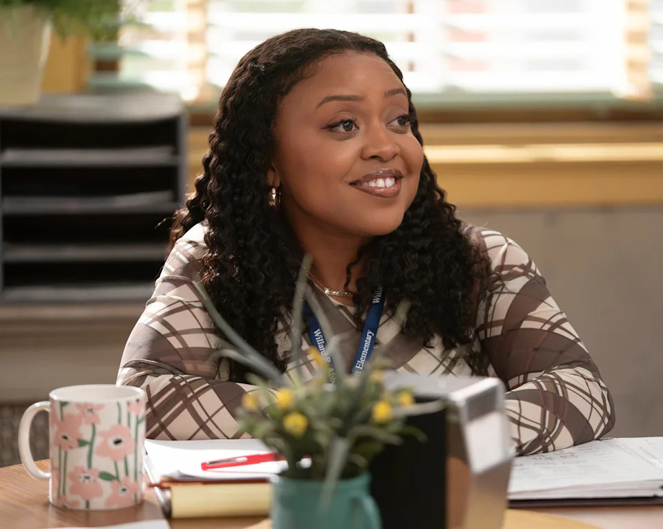 Person sitting at a desk with papers and a plant, smiling in a classroom setting, dressed in checkered clothing
