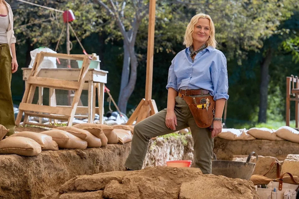 A person on an archaeological dig site, wearing casual work attire with a utility belt, stands confidently with one foot on a dirt mound