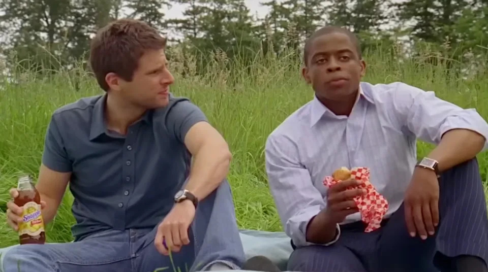 Shawn Spencer (James Roday) sits next to Gus Guster (Dulé Hill) on grass; one holds a drink and the other a wrapped sandwich, both gazing ahead thoughtfully