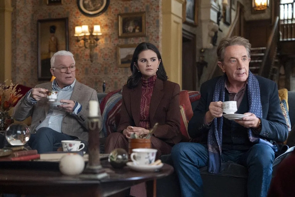 Three actors sit together, each holding a cup, on a cozy set decorated with vintage items, suggesting a scene from a TV show or movie
