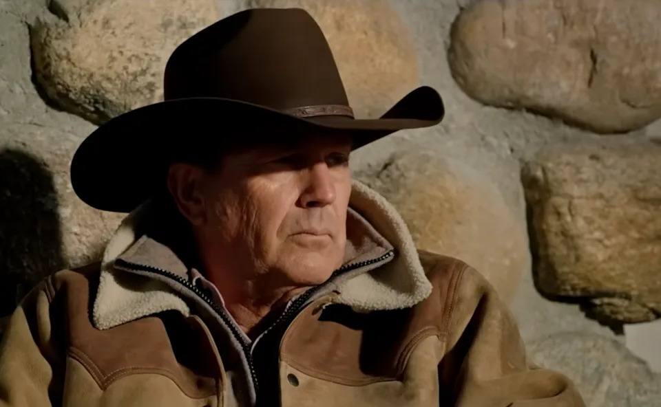 John Dutton (Kevin Cosner) in cowboy hat and jacket sitting by a stone wall, looking thoughtful