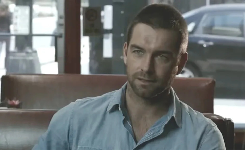 Lucas Hood (Anthony Starr) sits in a cafe, wearing a button-up shirt and looking slightly to his right