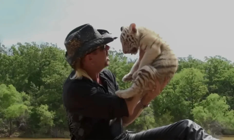 Joe Exotic in a cowboy hat, holding a white tiger cub, with trees in the background