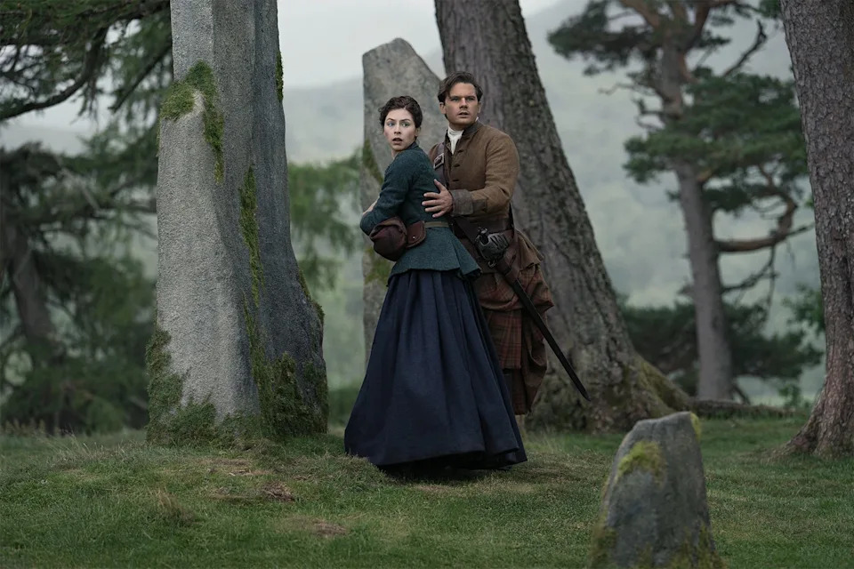 A man and woman in period costumes stand in a forest clearing with stone structures, looking alert in a dramatic scene