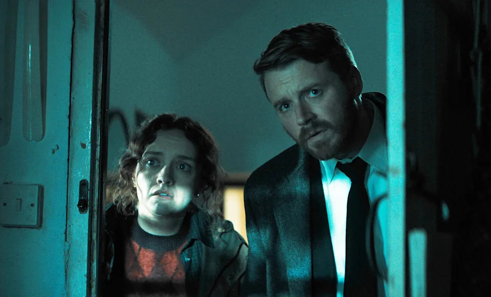 Two people stand in a dimly lit room, looking concerned. The man wears a suit and tie, while the woman is casually dressed