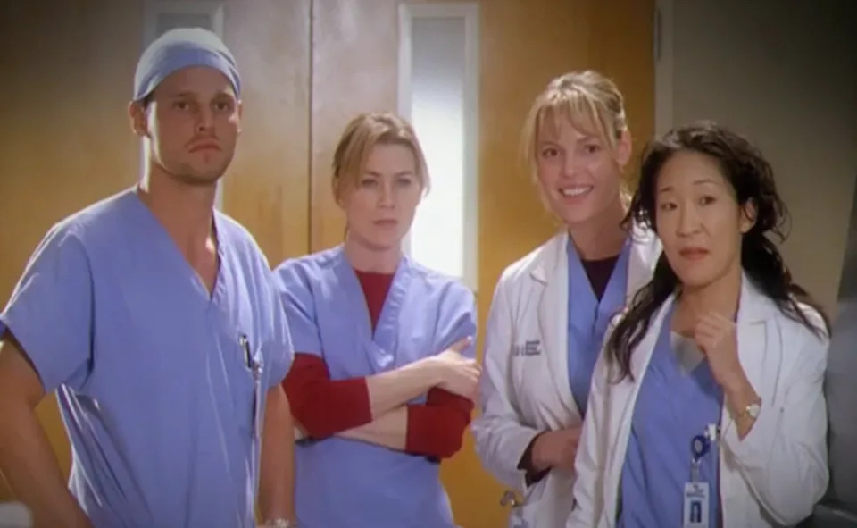 Alex Karev (Justin Chambers), Meredith Grey (Ellen Pompeo), Izzie Stevens (Katherine Heigl) and Cristina Yang (Sandrah Oh) wearing medical scrubs and lab coats stand in a hospital setting, appearing focused and attentive