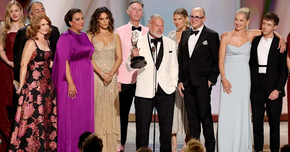 A group of people on stage at an awards ceremony, one holding an Emmy. They are dressed in formal attire, smiling and celebrating.