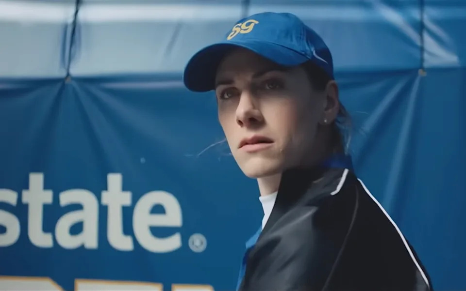 A focused athlete wearing a blue cap and a black jacket, standing in front of a blue backdrop with text.