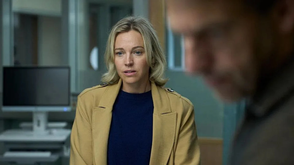 Loes Haverkort stands in a room, wearing a beige jacket over a blue shirt, looking intently at another person.