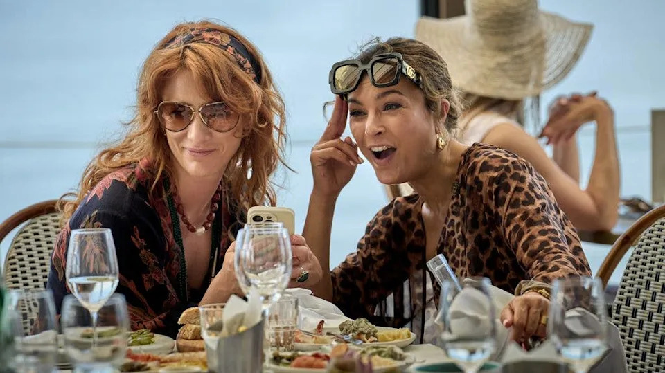 Two women at a dining table, one with sunglasses and leopard print top, the other with a headband and floral shirt, both smiling and looking at a phone.