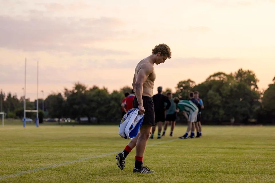 Athlete in black shorts and red socks holds a blue and white bag on a field with other players.