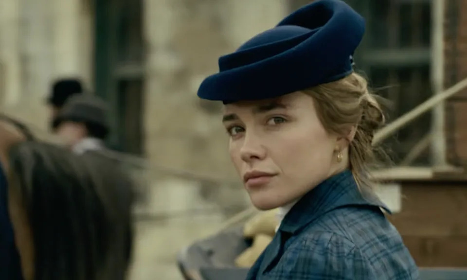 Florence Pugh in a blue hat and plaid coat stands near a horse carriage.